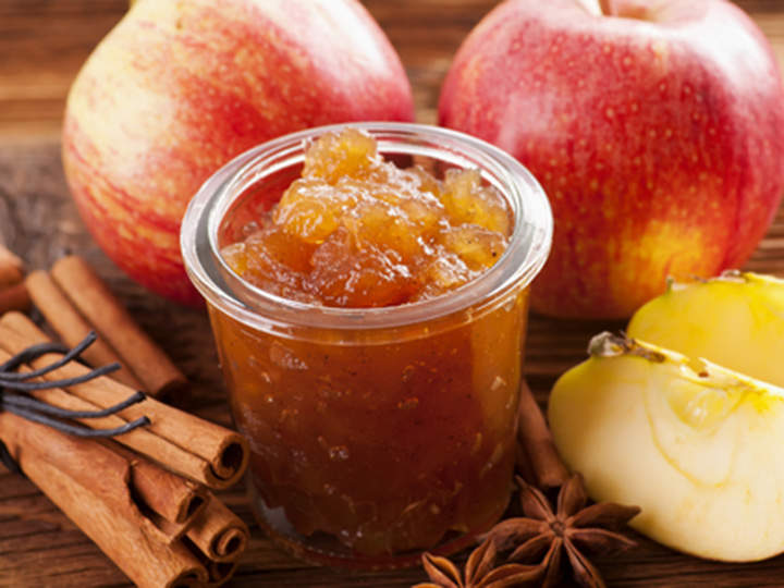 Ein Glas Apfelmarmelade, umgeben von frischen Äpfeln, Zimtstangen und Sternanis auf einer Holzoberfläche. Die Marmelade sieht stückig aus und hat eine goldbraune Farbe, die ein warmes Herbstthema hervorruft.