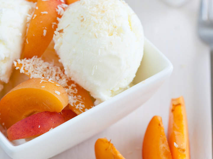 Eine weiße Schale gefüllt mit cremigem Vanilleeis, garniert mit Kokosraspeln, umgeben von Pfirsich- und Aprikosenscheiben auf hellem Untergrund. Im Vordergrund sind weitere Fruchtscheiben zu sehen.