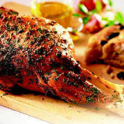 Eine gebratene Lammkeule, garniert mit Kräutern, auf einem Holzschneidebrett. Im Hintergrund befinden sich ein Stück Brot und ein kleines Glasgefäß mit Olivenöl, was die rustikale Präsentation noch verstärkt.