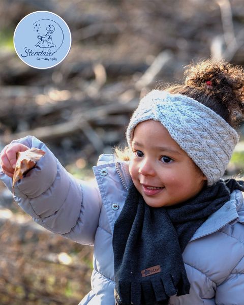 Un jeune enfant avec un manteau gris clair, une écharpe grise et un bandeau tricoté tient une feuille à l'extérieur. L'arrière-plan est flou à cause des branches et de la lumière du soleil. Dans le coin, on peut voir un logo rond "Sterntaler Allemagne 1965".