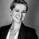 Une personne souriante aux cheveux courts, portant des boucles d'oreilles ornées et un élégant blazer noir sur un chemisier blanc, se tient devant un fond texturé. L'image est en noir et blanc.