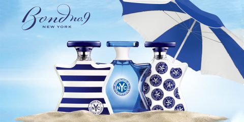 Trois flacons de parfum Bond No. 9 New York sont posés sur le sable devant un ciel bleu. Les flacons sont ornés de rayures et de motifs. À l'arrière-plan, un grand parasol bleu et blanc est déployé.