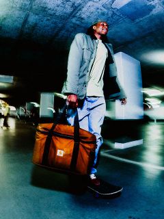 Homme faisant du skateboard dans un parking, portant un grand sac Carhartt marron, une veste verte et un jean sous une lumière artificielle.