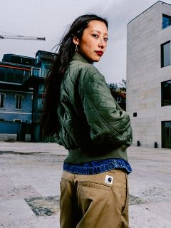 Une femme aux longs cheveux foncés, vêtue d'une veste matelassée verte et d'un pantalon marron clair, se tient à l'extérieur dans un environnement urbain et regarde par-dessus son épaule.