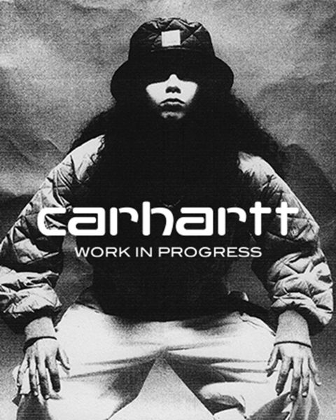 Image en noir et blanc d'une personne portant un chapeau et une veste, accroupie avec les mains sur les genoux. Le texte "Carhartt Work In Progress" est clairement visible au centre.