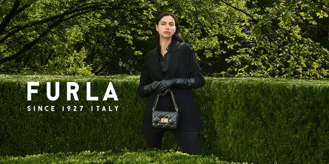 Une femme se tient devant une végétation luxuriante. Elle porte une tenue noire et des gants, et tient un sac à main noir matelassé. À gauche, on peut lire le texte « Furla Depuis 1927 Italie ».