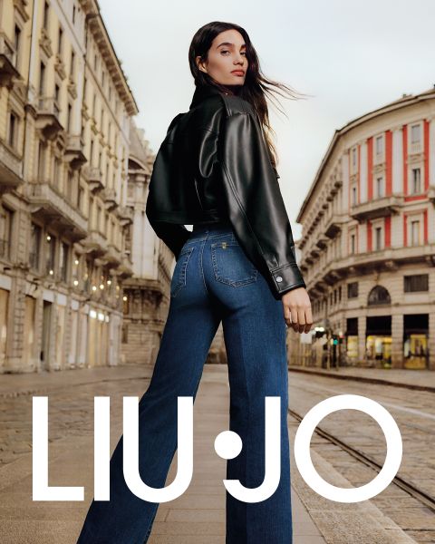 Une femme se tient dans une rue de la ville. Elle porte une veste en cuir noire et un jean bleu. Elle regarde par-dessus son épaule, tandis que ses longs cheveux flottent au vent. En arrière-plan, on aperçoit une architecture classique. En bas, on peut lire le texte « LIU JO ».