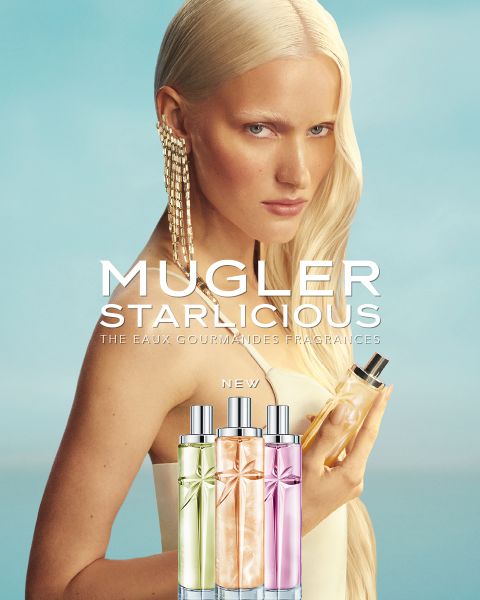 Une femme aux longs cheveux blonds tient un flacon de parfum dans sa main. Dans la partie inférieure de l'image, on peut voir trois flacons colorés « Mugler Starlcious » devant un ciel bleu.