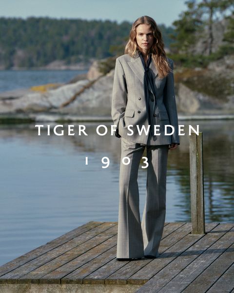 Une femme en tailleur gris se tient sur une jetée en bois au bord d'un lac aux rives rocheuses et boisées. Le texte « TIGER OF SWEDEN 1903 » est écrit en travers de l'image.