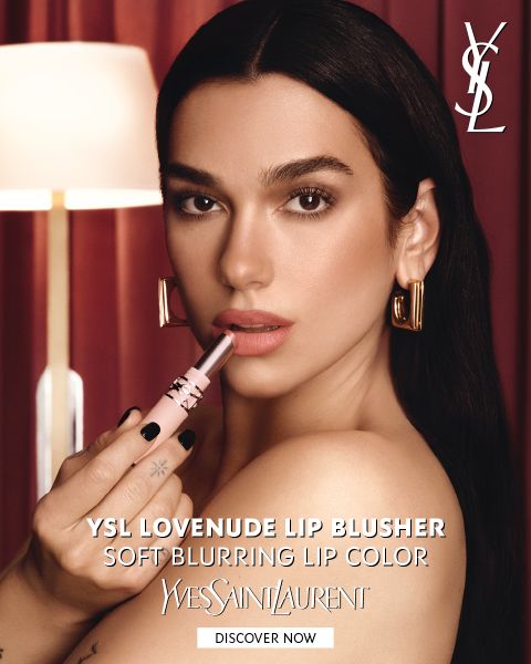 Une femme qui applique du rouge à lèvres rose et porte des créoles dorées, devant des rideaux rouges. Le texte fait la promotion du « YSL Lovenuede Lip Blusher », un rouge à lèvres à effet flouté doux.