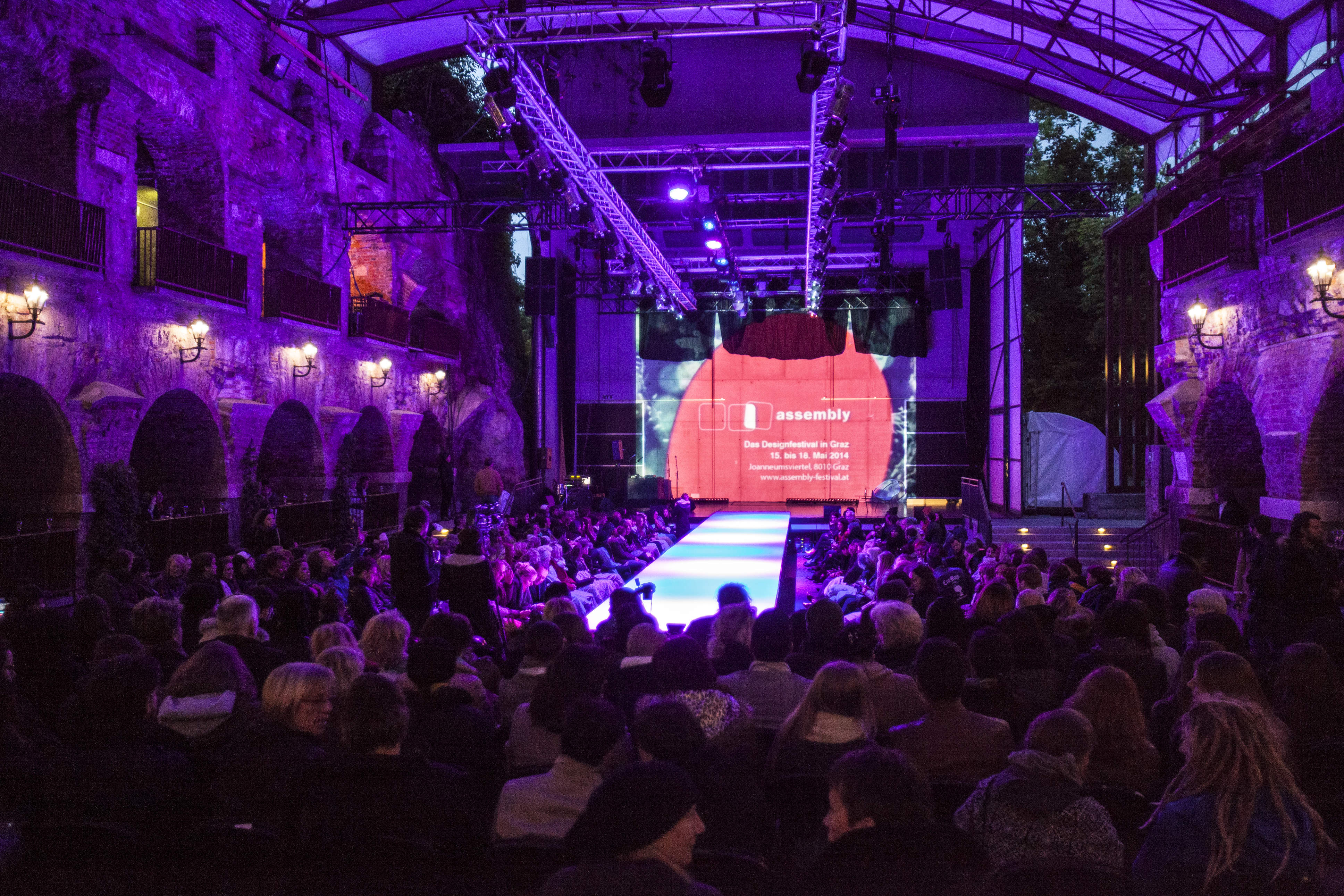 Eine schwach beleuchtete Modenschau mit einer Menschenmenge, die auf beiden Seiten sitzt. Der Hintergrund zeigt einen beleuchteten Text über einem roten Kreis. Der Veranstaltungsort hat Steinmauern und sanftes Licht, was eine intime Atmosphäre schafft.