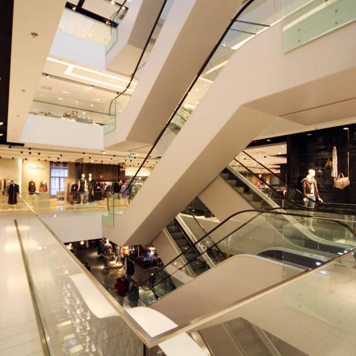 Ein modernes Einkaufszentrum mit mehreren Etagen und Rolltreppen. Der Raum ist gut beleuchtet und bietet Geschäfte mit Auslagen auf verschiedenen Ebenen. Überall sind Glasgeländer und elegante architektonische Designelemente zu sehen.