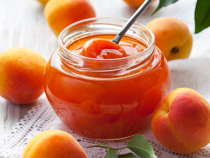 Ein Glas Aprikosenmarmelade mit einem Löffel steht auf einem Spitzentuch, umgeben von frischen Aprikosen. Die Marmelade ist glänzend und leuchtend orange und harmoniert mit den reifen Früchten daneben. Die Szene vermittelt ein Gefühl von Frische und hausgemachter Köstlichkeit.