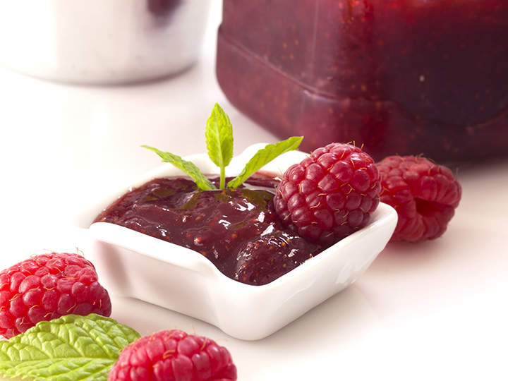 Ein kleines weißes Schälchen gefüllt mit Himbeermarmelade, garniert mit einem Zweig Minze. Frische Himbeeren und Minzblätter sind auf dem Schälchen verteilt. Im Hintergrund ist teilweise ein Glas Marmelade zu sehen.