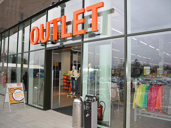 Ein Outlet-Store mit Glasfront und großem orangefarbenem „OUTLET“-Schild. Im Inneren sieht man Kleiderständer und Schaufensterpuppen. Ein Schild auf dem Bürgersteig wirbt für 20 % Rabatt.
