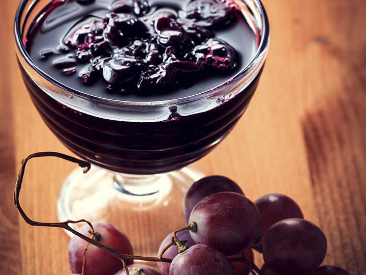 Auf einem Holztisch steht eine Glasschale mit dunkler Beerenmarmelade, neben einer kleinen Traube violetter Weintrauben. Die Beeren in der Marmelade glitzern und die Weintrauben haben ein natürliches, frisches Aussehen.