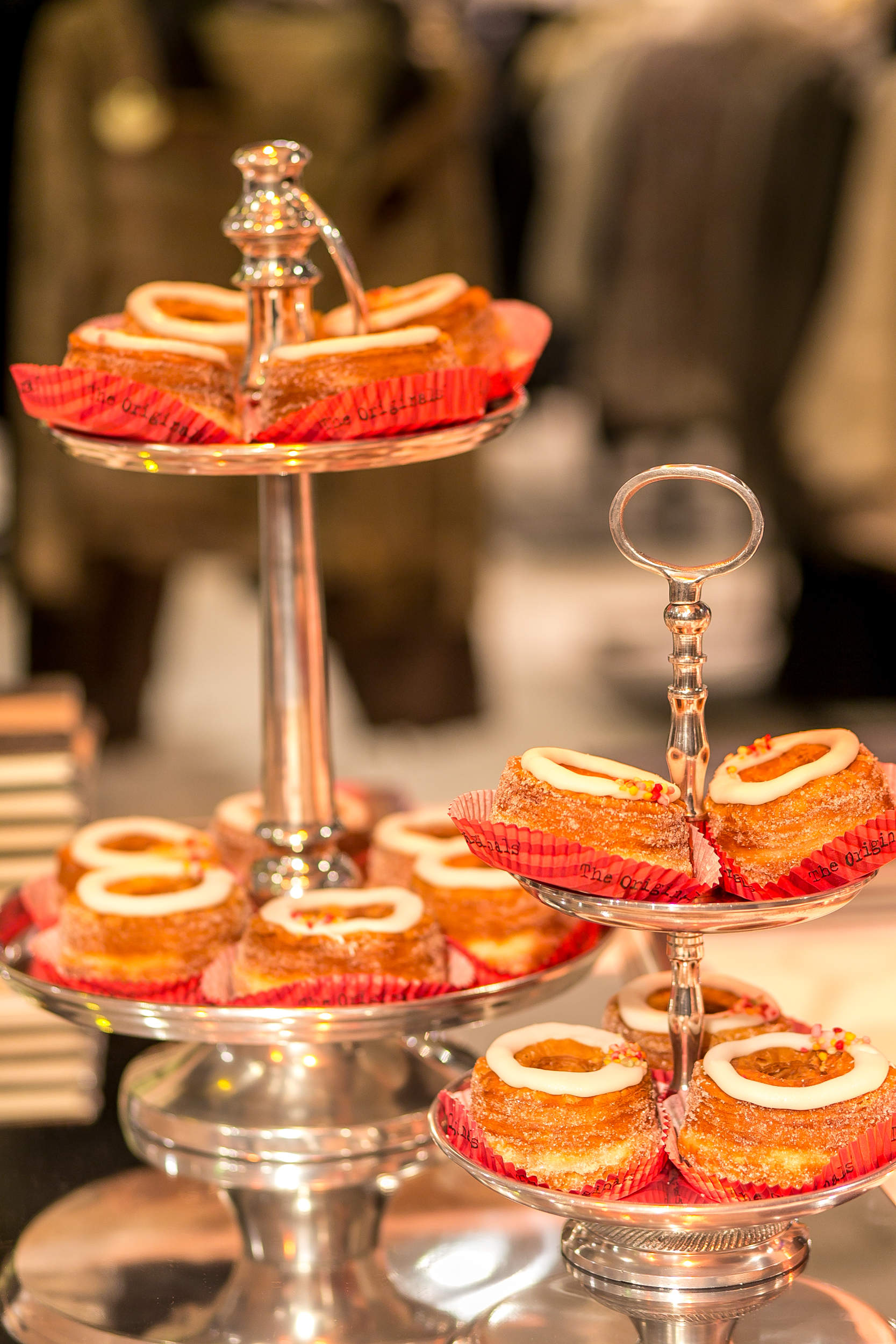 Auf einem mehrstöckigen silbernen Tablett sind mehrere kleine Kuchen mit weißem Zuckerguss und bunten Streuseln ausgestellt, die jeweils in einer roten Papierunterlage liegen. Der Hintergrund ist verschwommen, was die elegante Präsentation der Desserts unterstreicht.