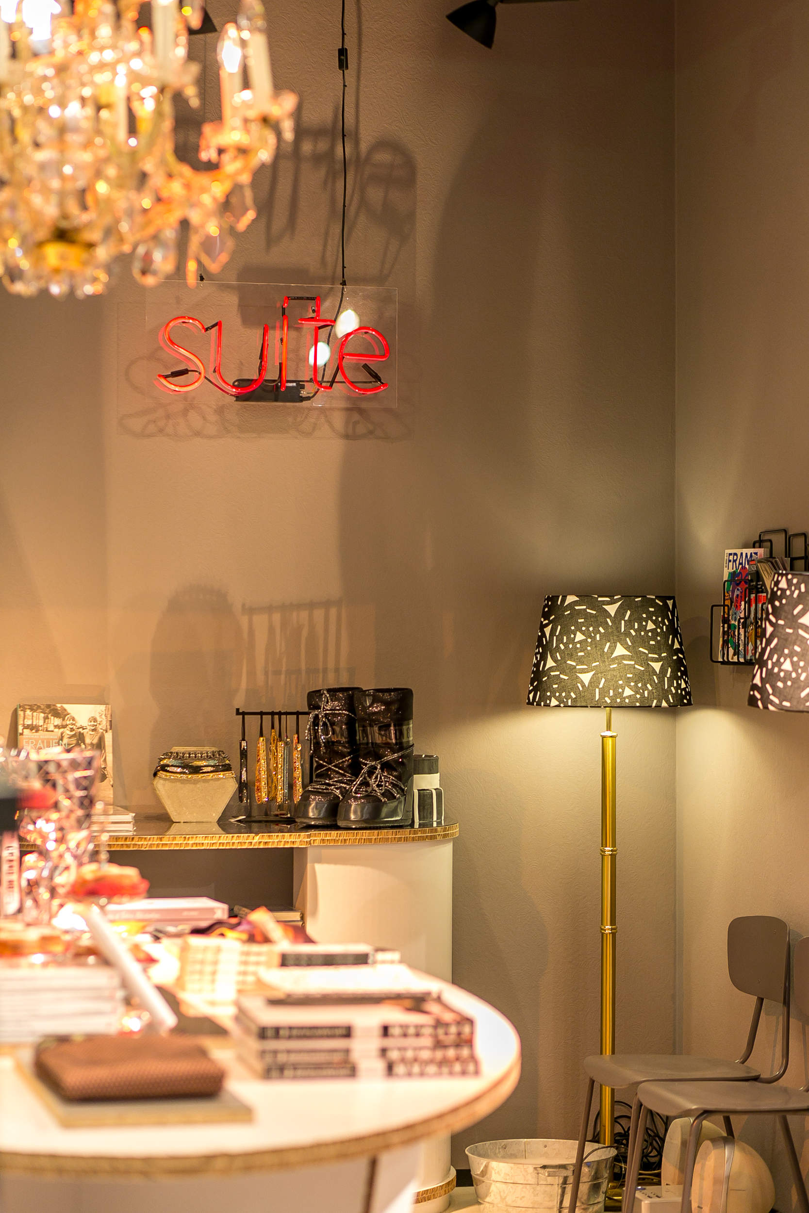 Ein gemütliches Boutique-Interieur mit einem Kronleuchter, einem Neonschild mit der Aufschrift „Suite“, Dekorationsgegenständen auf Regalen, Zeitschriften auf einem Ständer und einer gemusterten Stehlampe neben einem grauen Stuhl. Die Wände sind in einem warmen Beigeton gehalten, was zur einladenden Atmosphäre beiträgt.