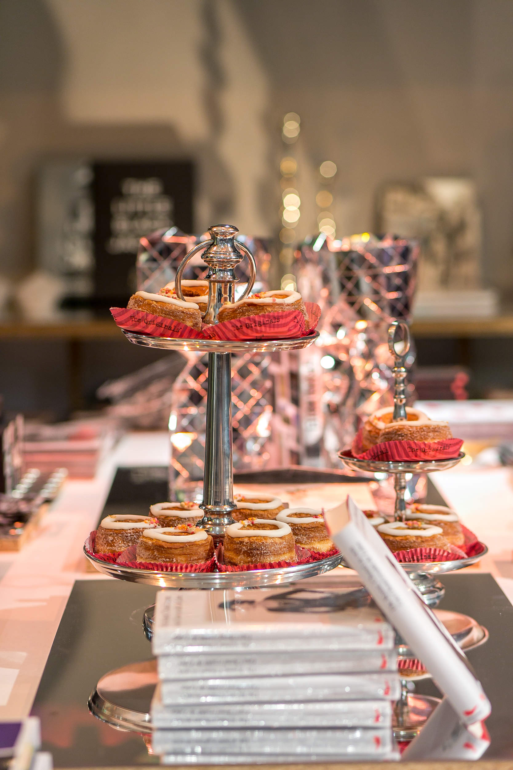 Auf einem zweistöckigen silbernen Dessertständer stehen ordentlich angeordnete Macarons in rosa Verpackungen auf einem Tisch. Im Hintergrund sind verschiedene Bücher und Dekoartikel verschwommen und schaffen eine gemütliche, gehobene Atmosphäre.