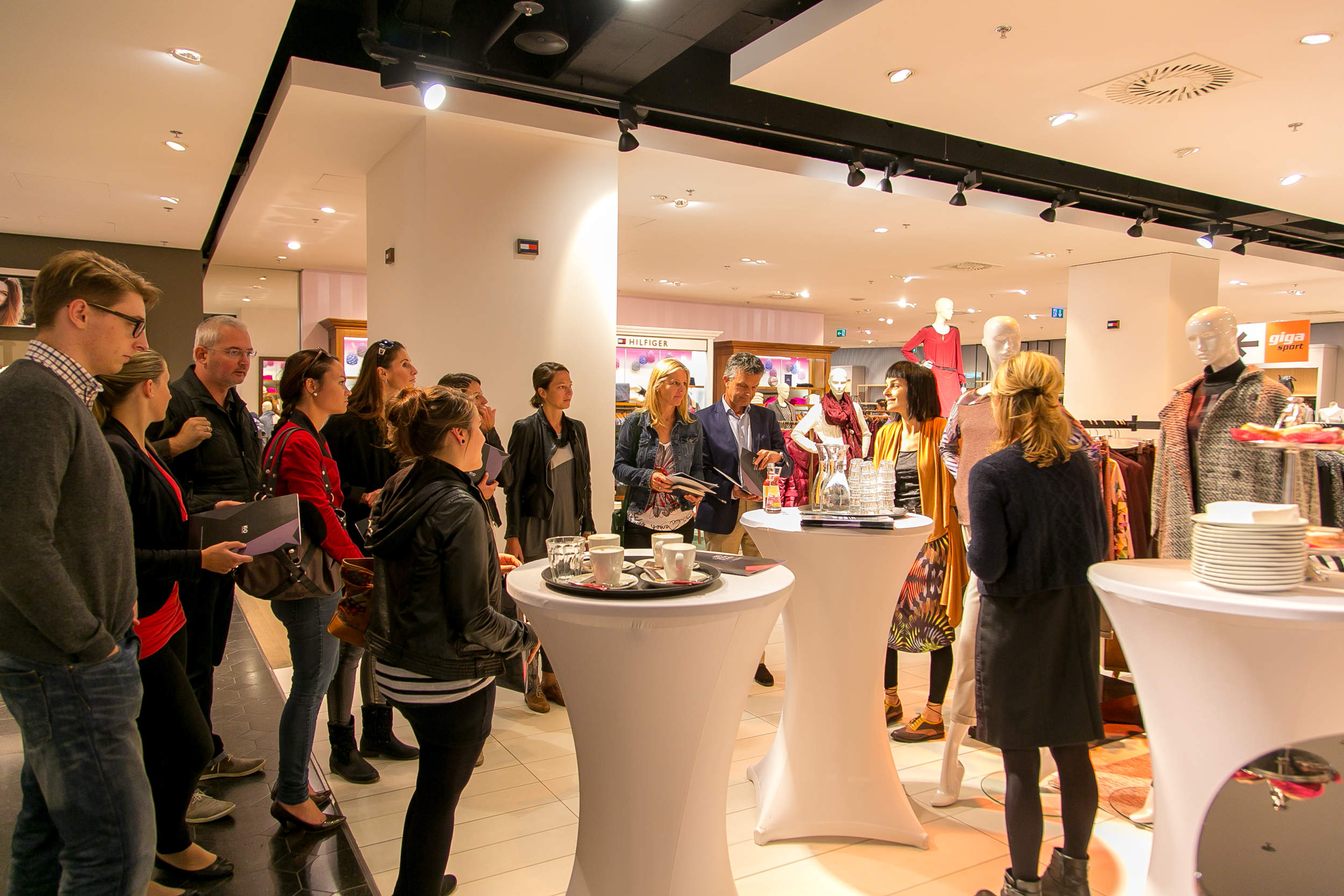 Eine Gruppe von Menschen versammelt sich um zwei hohe Tische in einem gut beleuchteten Bekleidungsgeschäft. Sie sind in eine Diskussion oder Präsentation vertieft, im Hintergrund präsentieren Schaufensterpuppen Outfits. Auf einigen Tischen stehen Teller und Tassen.