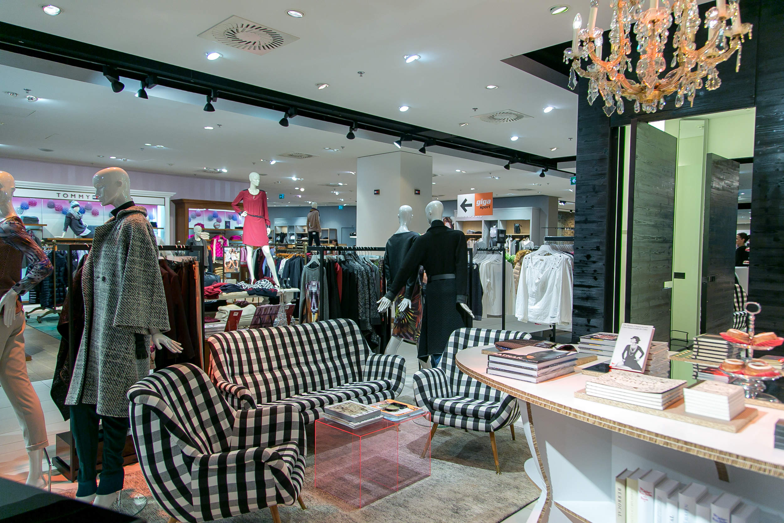 Ein stilvolles Interieur eines Bekleidungsgeschäfts mit Schaufensterpuppen, die verschiedene Outfits präsentieren. Der Raum umfasst einen Sitzbereich mit einem schwarz-weiß gestreiften Sofa, einem Couchtisch und dekorativen Büchern. Schicke Beleuchtung und modernes Dekor runden die Szene ab.