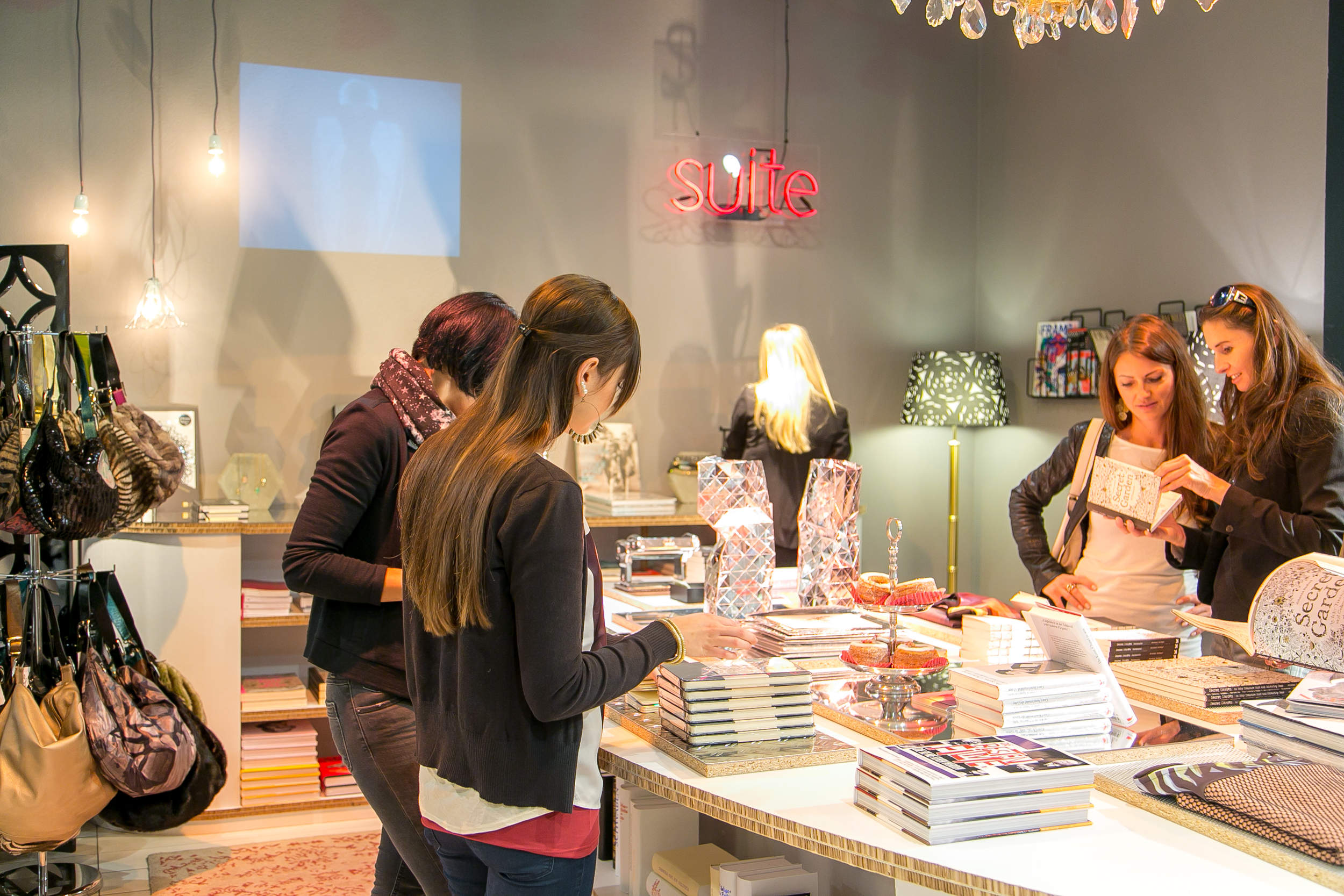 Eine Gruppe von Frauen geht in einem hell erleuchteten Geschäft einkaufen. Sie stöbern in Büchern und Dekorationsartikeln, die auf einem großen Tisch ausgestellt sind. An den Wänden sind Handtaschen und weitere Dekorationen zu sehen. Im Hintergrund leuchtet ein Neonschild mit der Aufschrift „Suite“.