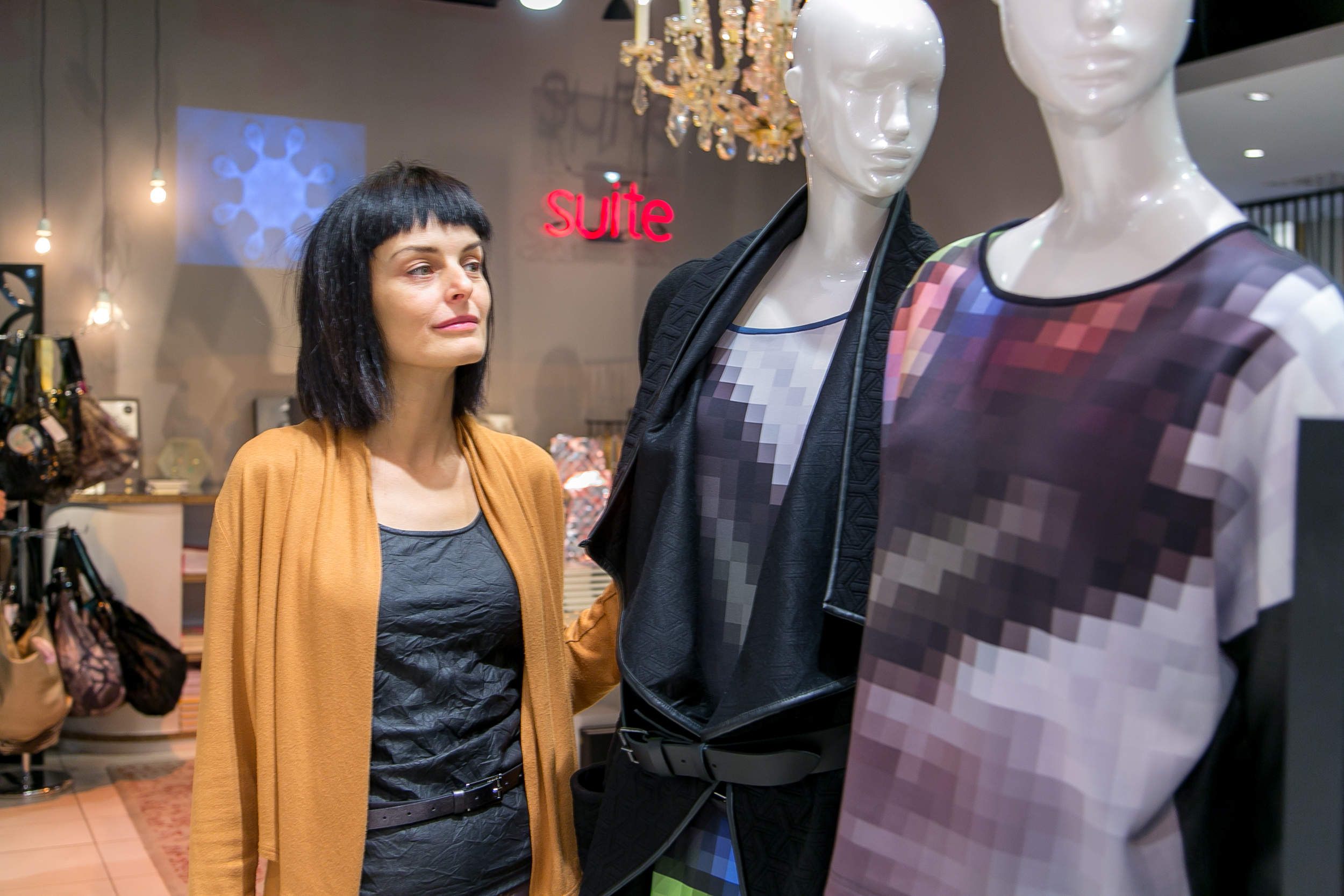 Eine Frau mit dunklem Haar und einer orangefarbenen Strickjacke steht neben zwei Schaufensterpuppen in verpixelten Outfits in einer Modeboutique. Im Hintergrund sind ein Kronleuchter und verschiedene Handtaschen zu sehen.