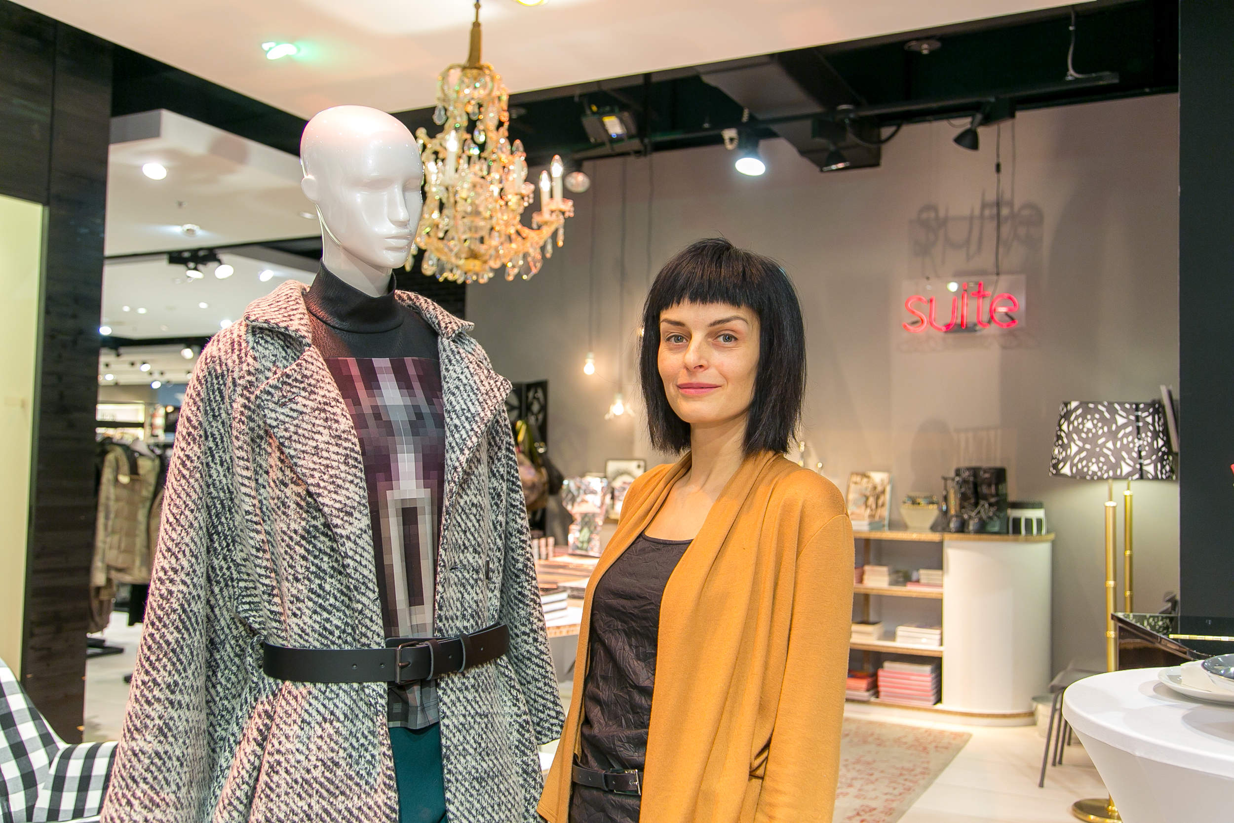 Eine Frau mit Kurzhaarschnitt steht in einer Boutique neben einer Schaufensterpuppe, die einen gemusterten Mantel trägt. Der Laden ist stilvoll, mit Kronleuchter und modernem Dekor, im Hintergrund steht ein Neonschild mit der Aufschrift „Suite“.