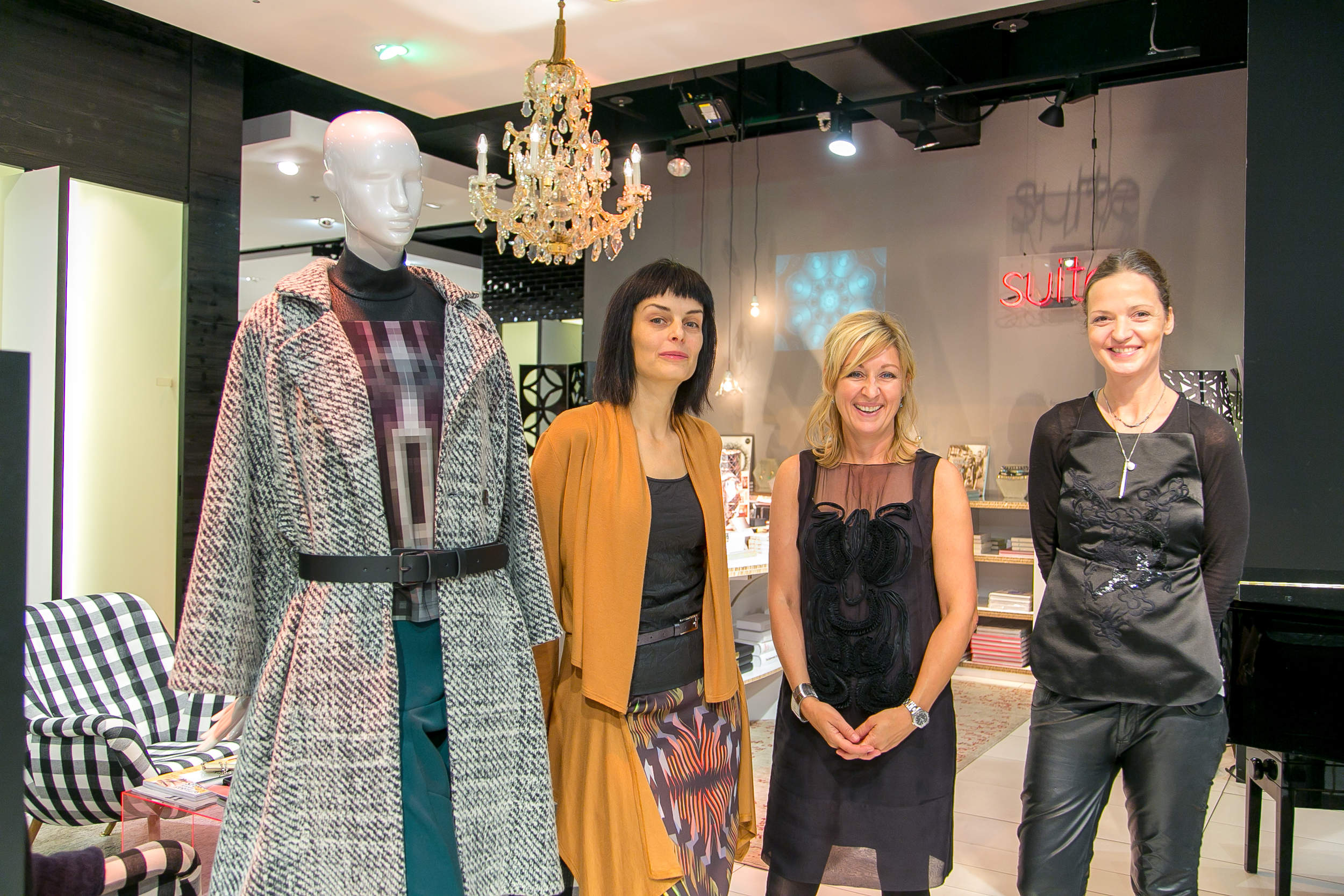 Drei Frauen stehen lächelnd neben einer Schaufensterpuppe in einem stylischen Outfit unter einem Kronleuchter in einem modernen Ladenambiente.