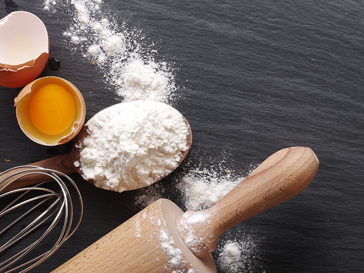 Ein hölzernes Nudelholz, ein Schneebesen, ein aufgeschlagenes Ei mit Eigelb und ein Holzlöffel mit Mehl sind auf einer dunklen Schieferplatte angeordnet. Überall ist Mehl verstreut, sodass eine rustikale Backszene entsteht.