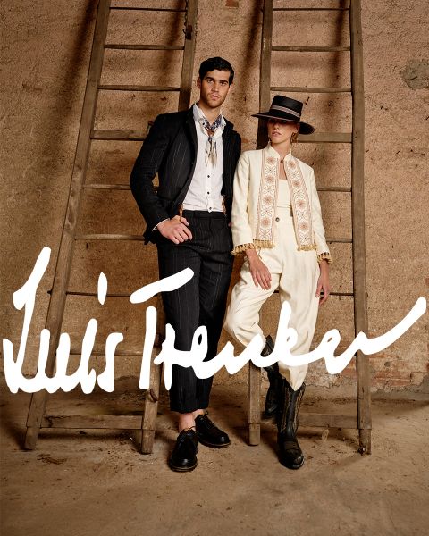 Un homme et une femme portant des tenues élégantes d'inspiration vintage se tiennent entre deux échelles en bois devant un mur rustique. L'homme porte un costume à rayures foncées, la femme un costume couleur crème avec un chapeau et des bottes. « Luis Trenker » est écrit en gras dessus.