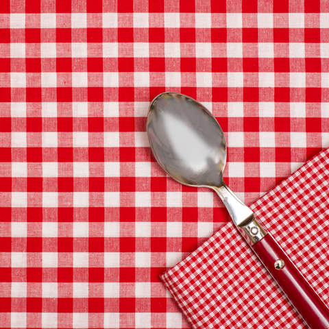 Ein Metalllöffel mit rotem Griff liegt auf einer rot-weiß karierten Tischdecke. Daneben liegt eine passende Serviette.
