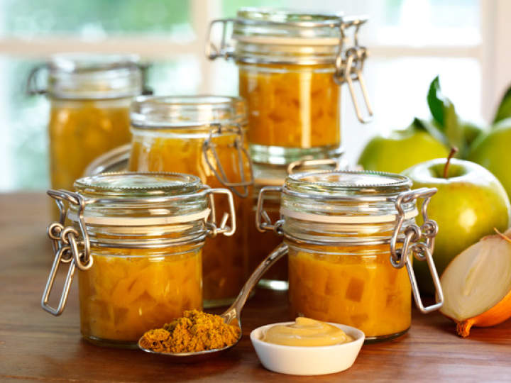 Gläser mit selbstgemachtem Chutney stehen auf einem Holztisch, umgeben von grünen Äpfeln. Ein Löffel mit Gewürzen und ein Schälchen Senf runden das kulinarische Ambiente ab. Die Gläser sind mit Metallclips verschlossen und durch ein Fenster im Hintergrund fällt Sonnenlicht.