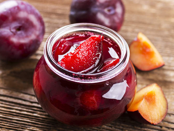 Auf einem Holztisch steht ein Glas mit Pflaumenmarmelade. Ein paar ganze Pflaumen und geschnittene Pflaumenstücke liegen verstreut herum. Die Marmelade sieht dick und farbintensiver aus.