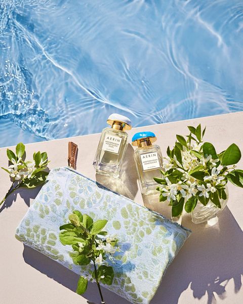 Deux flacons de parfum, une pochette ornée de fleurs et de petits vases avec des fleurs blanches sont disposés sur une tablette au bord de la piscine, près de l'eau bleue claire.