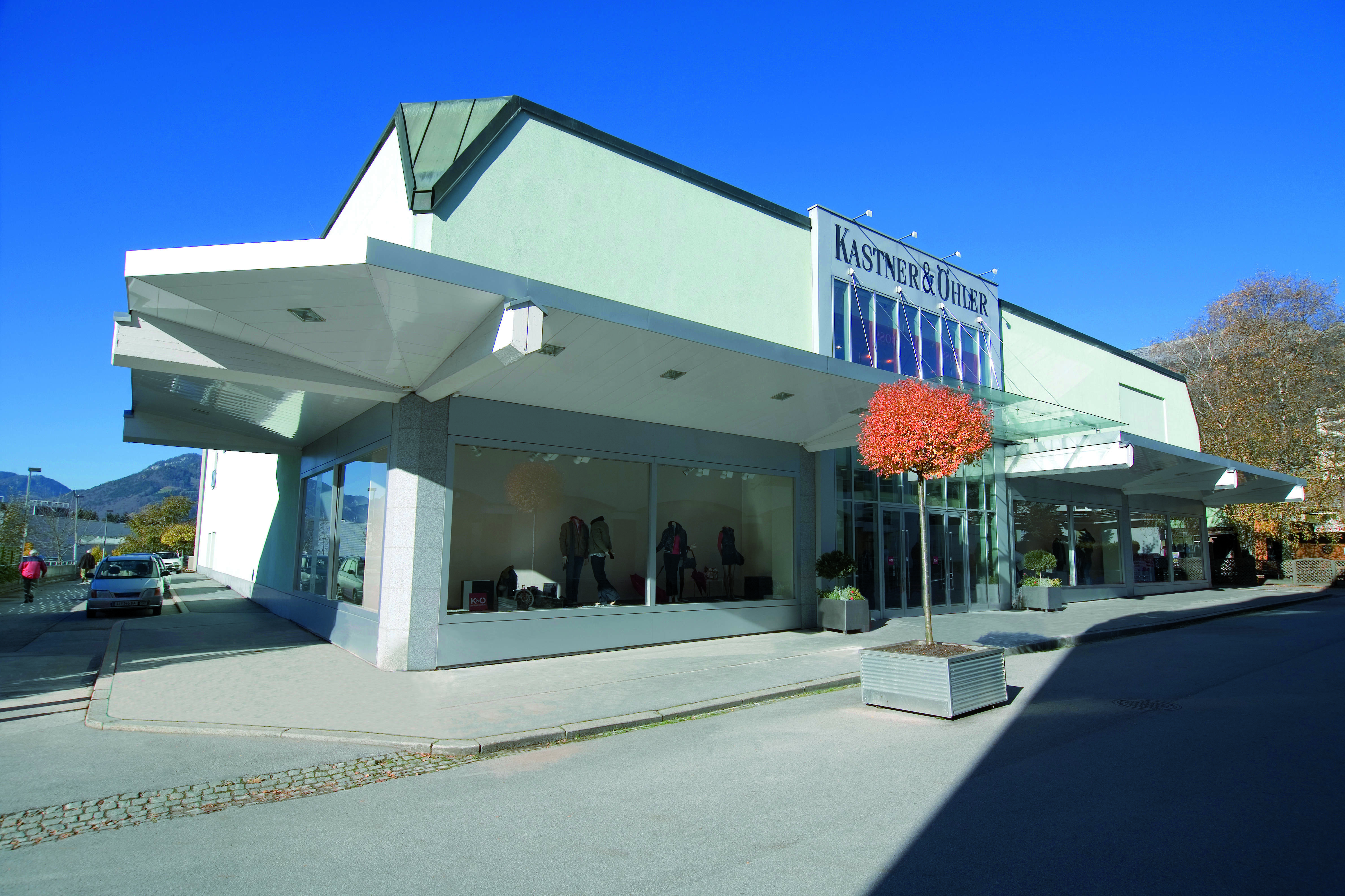Ein modernes Geschäftsgebäude mit einer Glasfront, in der Kleidung ausgestellt ist. Das Gebäude zeichnet sich durch ein elegantes Design mit schrägen Dächern und großen Fenstern aus. Davor steht ein kleiner Baum mit rotem Laub und in der Nähe parkt unter einem klaren blauen Himmel ein Auto.