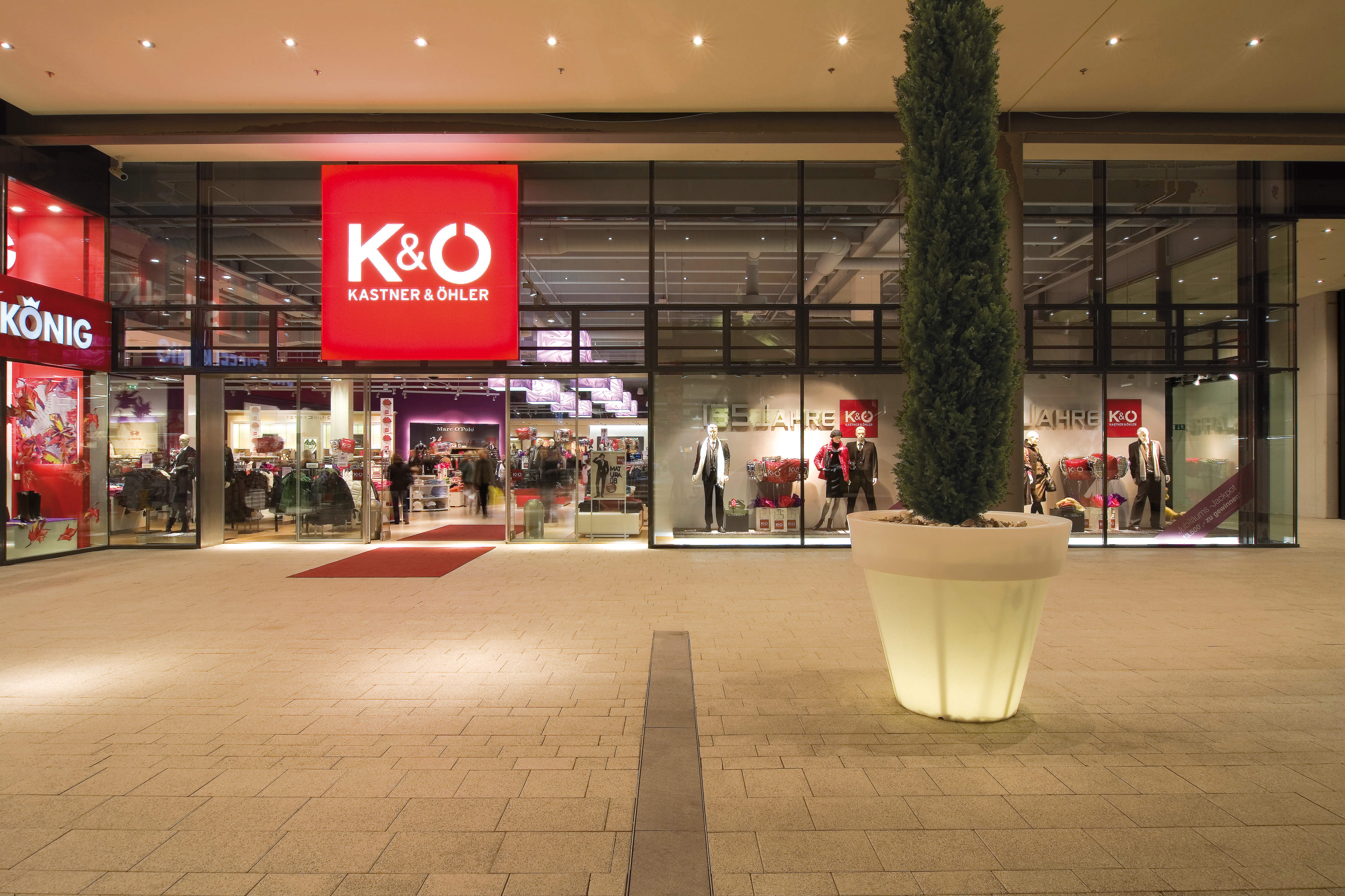 Schaufenster von K&O (Kastner & Öhler) mit großen Fenstern, in denen Schaufensterpuppen und Kleidung ausgestellt sind. Das rote Schild ist gut sichtbar über dem Eingang angebracht. Draußen steht auf einem gut beleuchteten gepflasterten Platz ein großer Topfbaum.