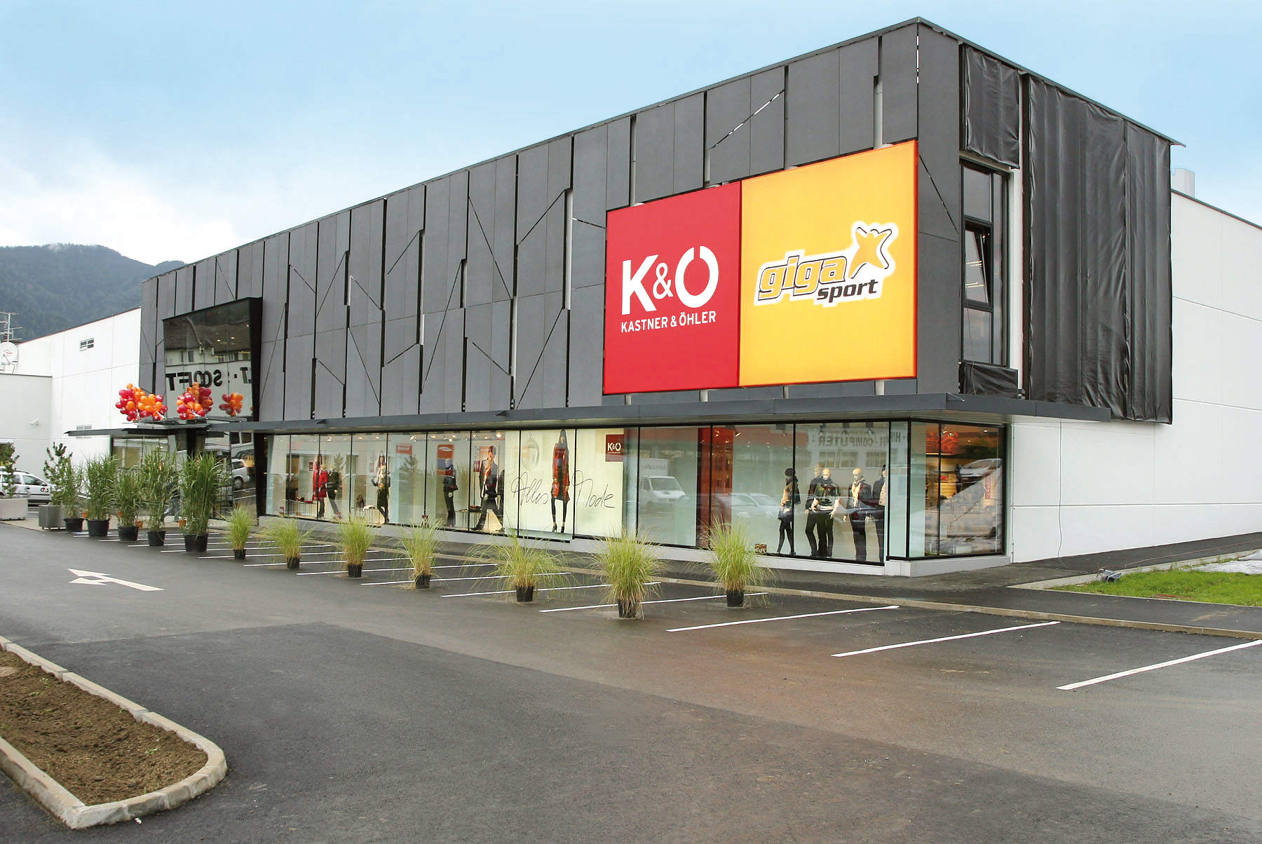 Ein modernes Geschäftshaus mit einem großen K&O- und Giga Sport-Logo an der Fassade. An den hohen Fenstern hängen Schaufensterpuppen, und am Eingang stehen Topfpflanzen. Der Himmel ist bedeckt.