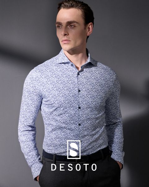 Un homme portant une chemise à manches longues à motifs et un pantalon foncé se tient devant un fond gris. Il regarde sur le côté, les mains dans les poches. L'inscription "DESOTO" et le logo DESOTO sont visibles en bas.
