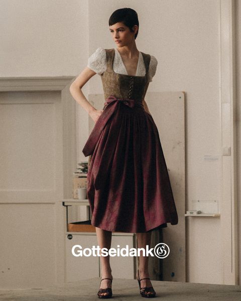 Une personne se tient à l'intérieur et porte une tenue traditionnelle bavaroise, composée d'un chemisier blanc, d'un gilet décoratif et d'une longue jupe violette avec un nœud. En arrière-plan, on aperçoit un mur nu et une pile de livres. En bas, on peut lire le mot « Gottseidank ».