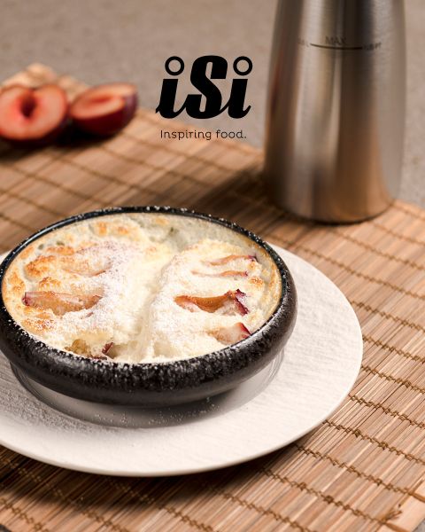 Un dessert cuit au four saupoudré de sucre glace est servi dans un bol noir, avec des prunes et un distributeur en métal en arrière-plan.