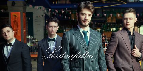Quatre hommes en costume formel posent avec assurance dans un cadre élégant avec une décoration suspendue. L'inscription "Seidenfalter" est affichée en travers de l'image.