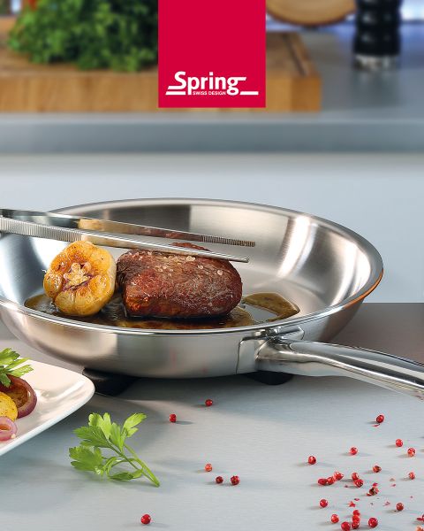 Sur un plan de travail de cuisine, une poêle en acier inoxydable contient un steak et de l'ail, que l'on retourne avec une pince. À côté se trouvent des grains de poivre rouge et du persil.