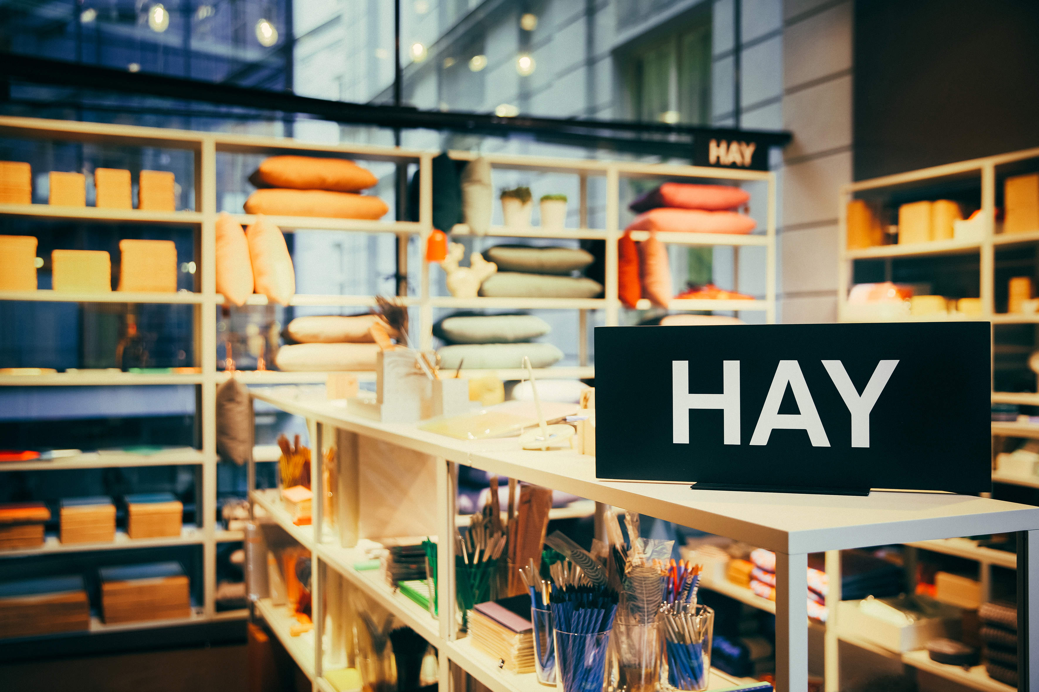 Ladeninneres mit Regalen voller bunter Kissen, ordentlich angeordneten Haushaltswaren und Schreibwaren. Vor den Regalen ist ein auffälliges schwarzes Schild mit der weißen Aufschrift „HAY“ angebracht.