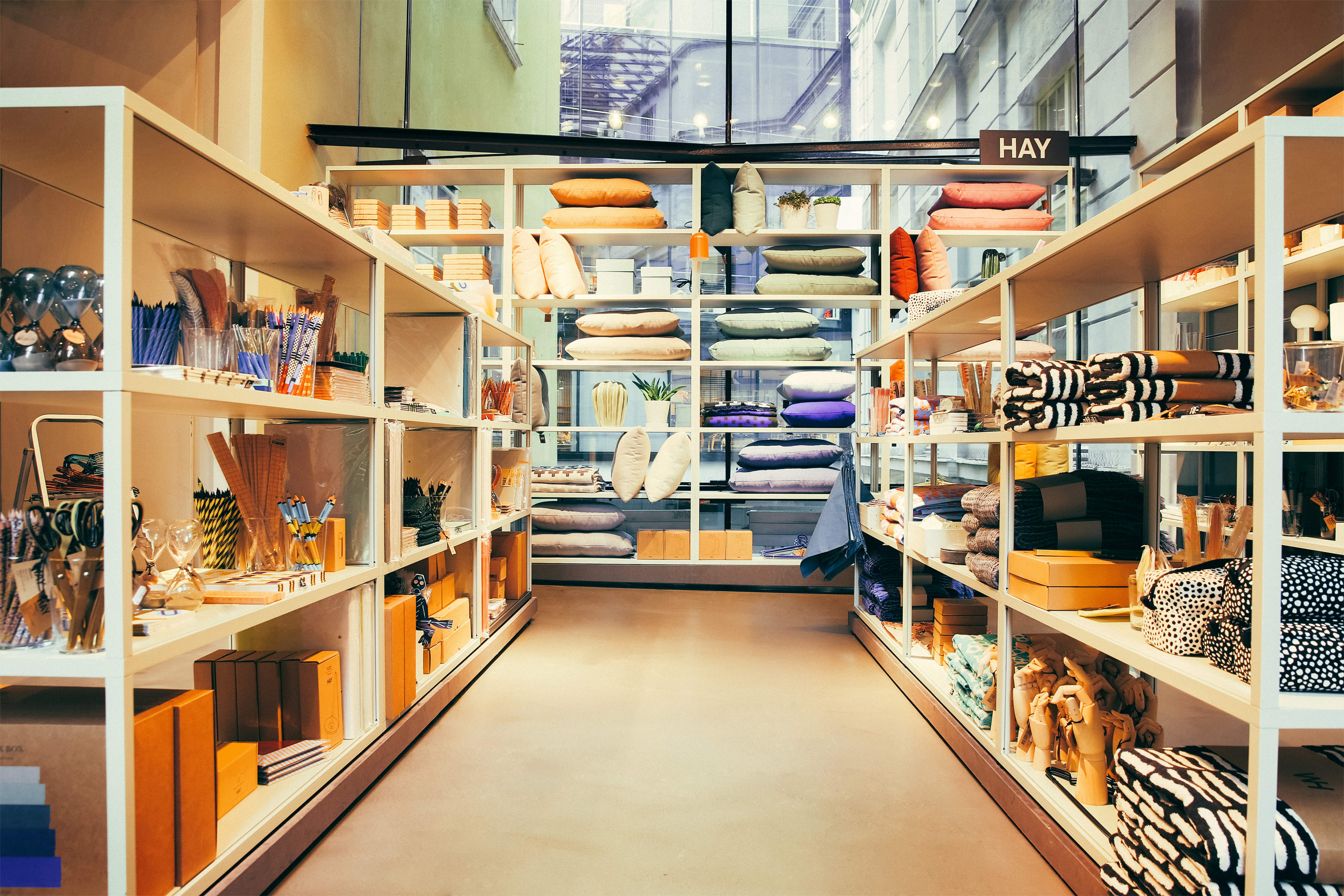 In einem gut organisierten Ladengang sind ordentlich gestapelte Kissen, Textilien und verschiedene moderne Wohnaccessoires auf weißen Regalen ausgestellt. Im Hintergrund sorgt ein großes Fenster für natürliches Licht.