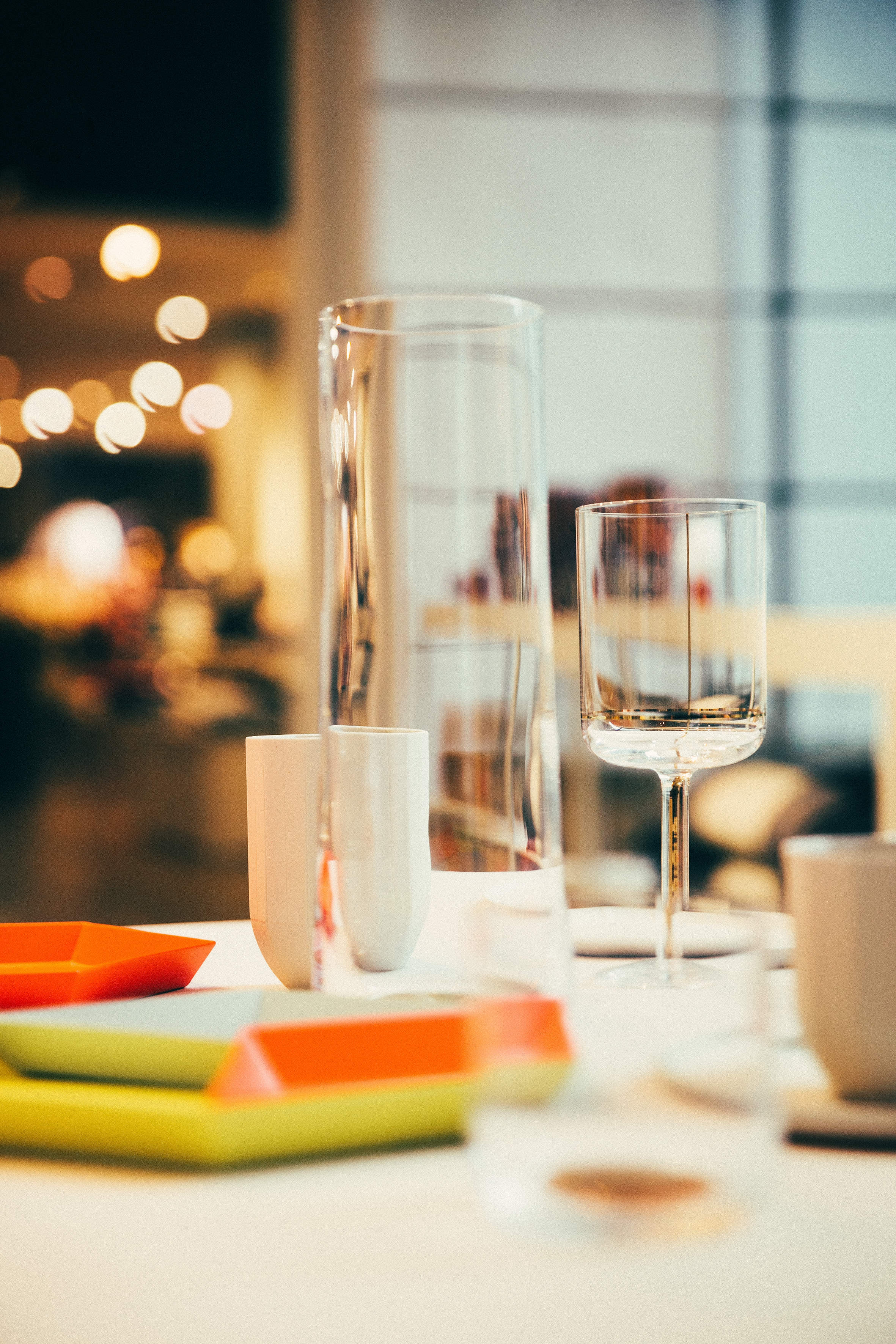 Auf einem schön gedeckten Tisch stehen ein klares Weinglas, eine hohe, schlanke Vase, bunte rechteckige Teller und eine kleine weiße Tasse. Der Hintergrund hat eine warme, sanft verschwommene Atmosphäre und runde Lichter unterstreichen die gemütliche Atmosphäre.