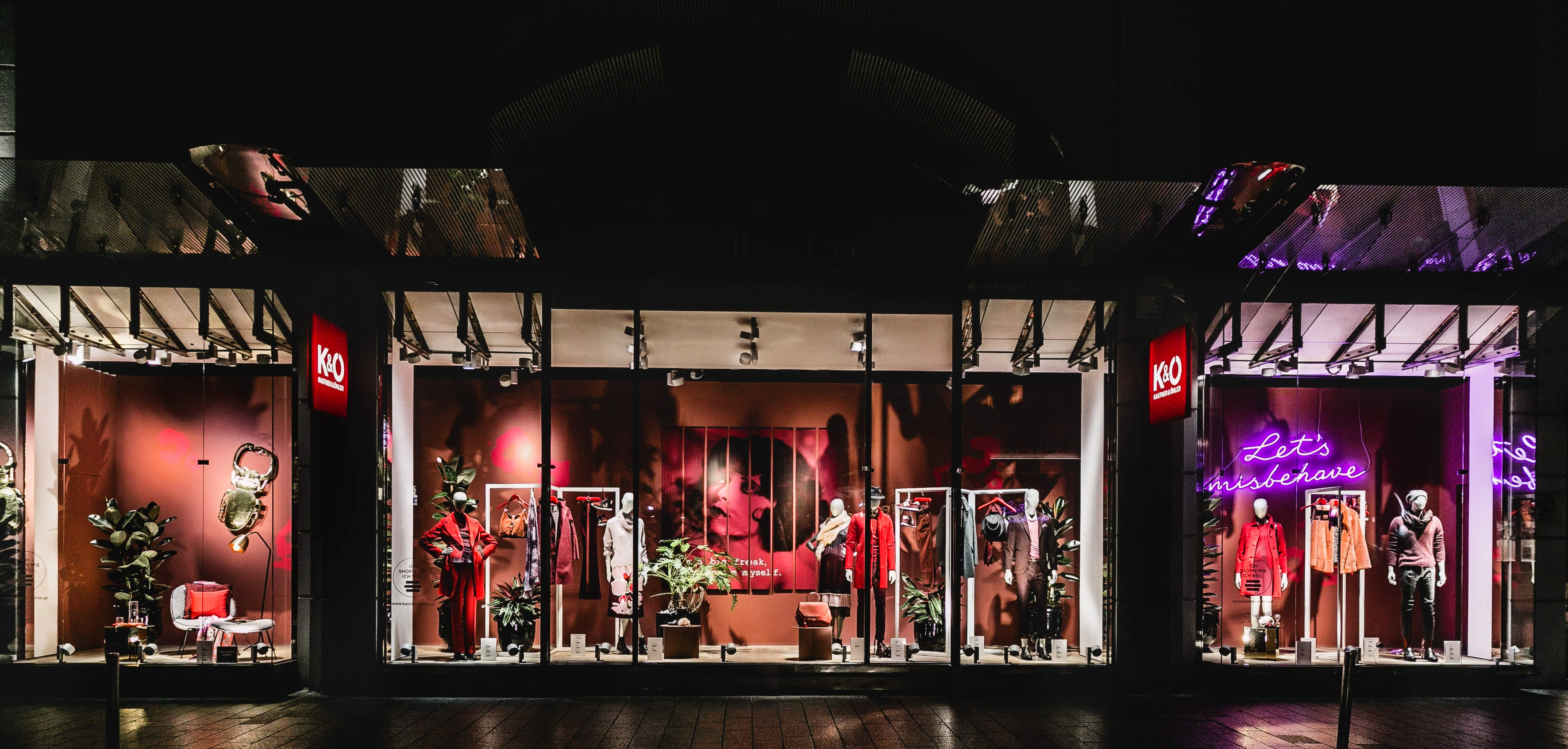 Ein stilvolles Schaufenster einer Boutique mit Schaufensterpuppen in schicken roten und schwarzen Outfits. Zur Dekoration gehören Pflanzen und ein Neonschild mit der Aufschrift „Lasst uns uns daneben benehmen“. Die beleuchteten Schilder verleihen der dunklen Außenfassade einen lebendigen Touch.