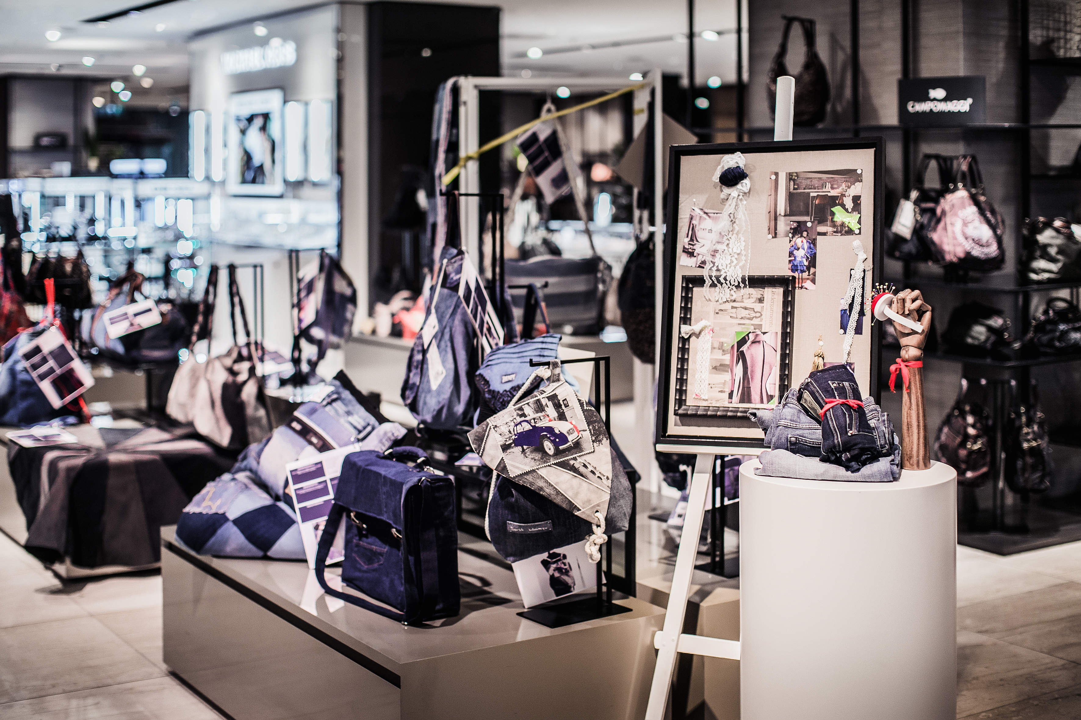 Eine moderne Ladenauslage mit verschiedenen Designertaschen in anspruchsvollem Ambiente. Im Vordergrund ist eine künstlerische Tafel mit Skizzen und Stoffmustern zu sehen. Im Hintergrund sind weitere Taschen und stilvolle Deko-Elemente auf Regalen zu sehen.