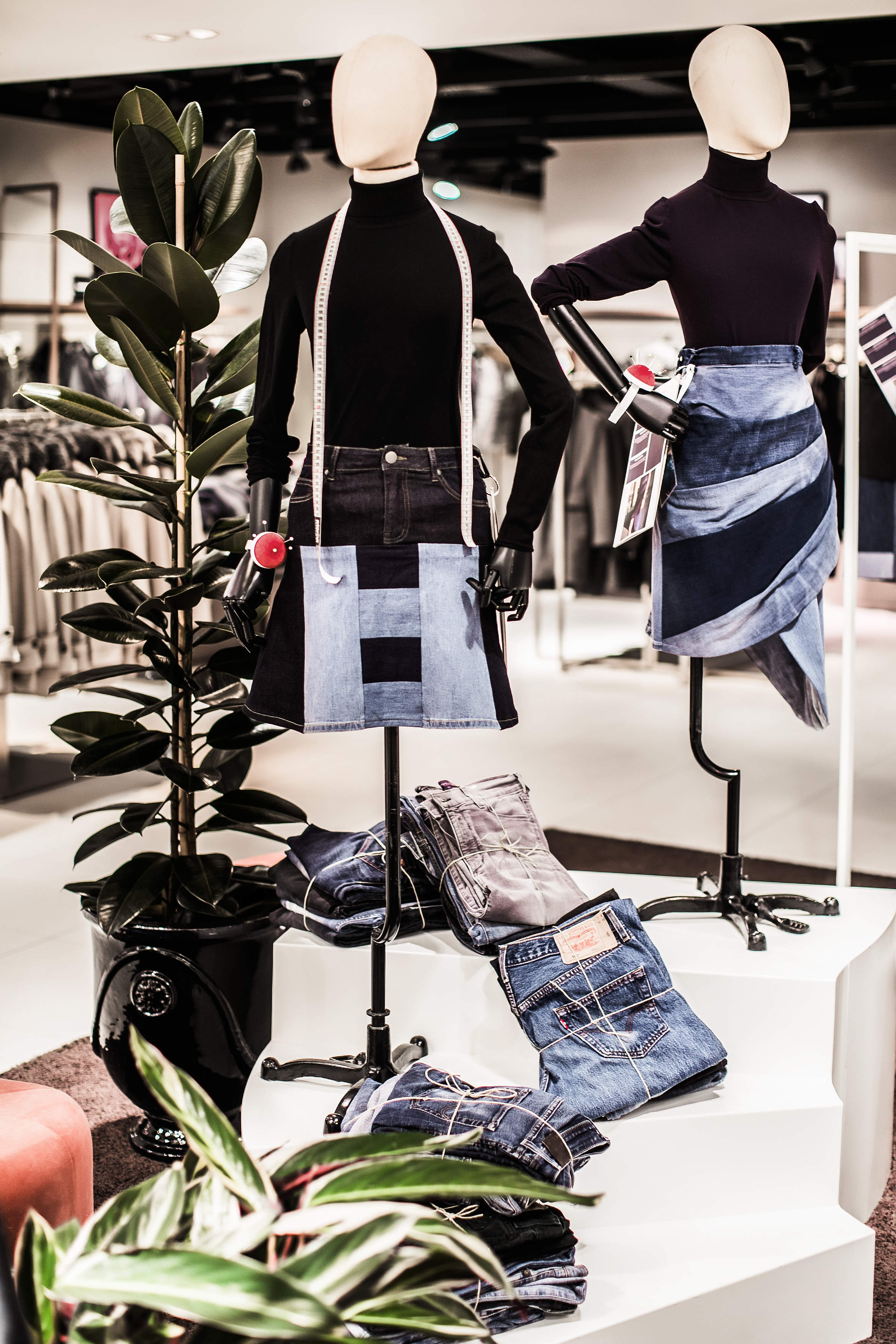 Zwei Schaufensterpuppen in Patchwork-Jeansröcken und schwarzen Oberteilen stehen auf einer Plattform in einem Bekleidungsgeschäft. Daneben steht eine Topfpflanze, und auf den Stufen sind gefaltete Jeans ausgestellt. Der Laden ist innen sanft beleuchtet.
