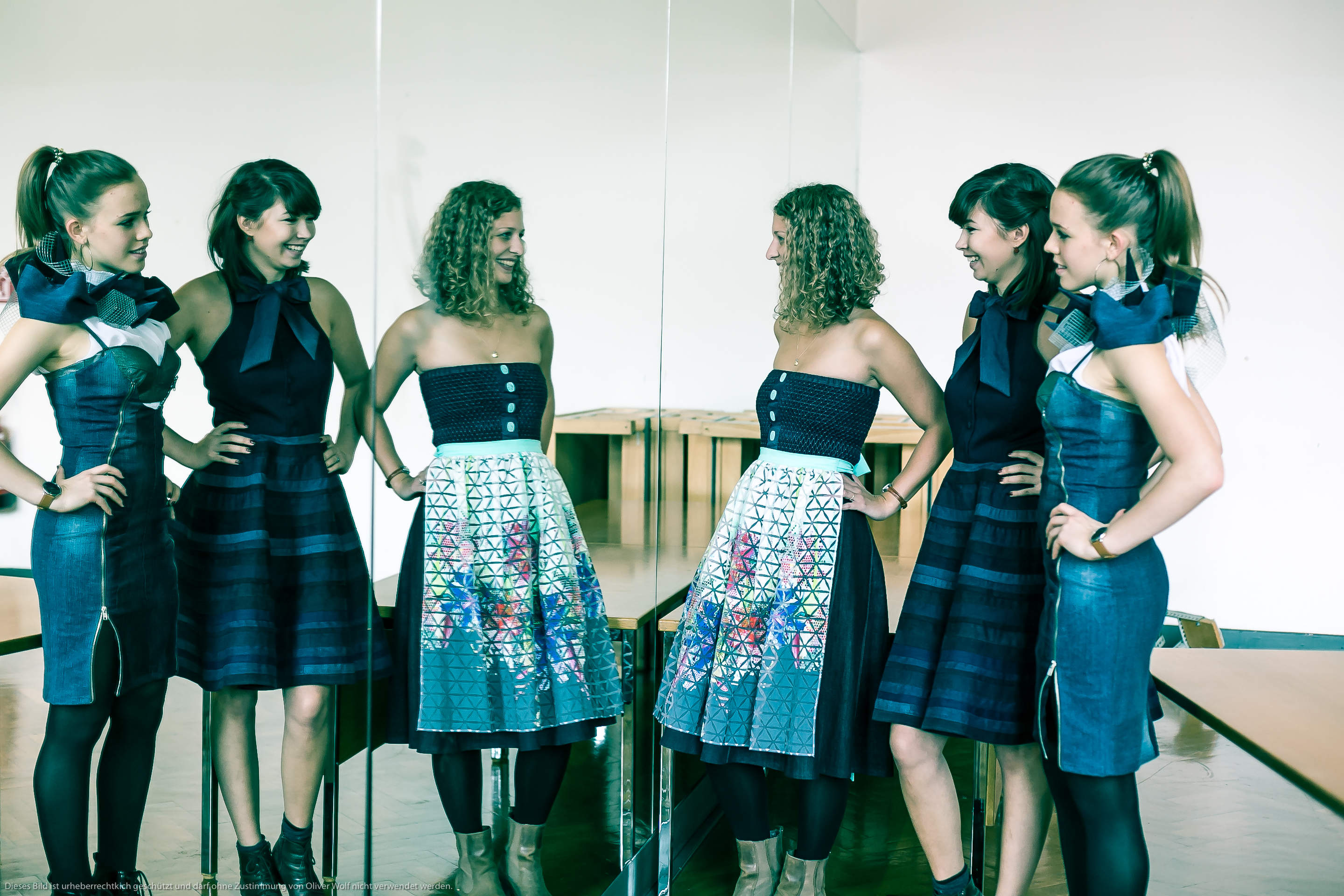 Zwei Frauen in passenden blauen Kleidern und schwarzen Leggings blicken in eine verspiegelte Wand, wodurch die Illusion von vier identischen Figuren entsteht. Sie stehen in einem hellen Raum mit Holzbänken, jede hat eine Hand auf der Hüfte und lächelt über die Spiegelungen.